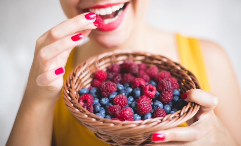 The young woman smiles while holding and eating from a basket filled with raspberries and blueberries