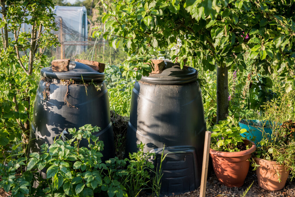 Two black plastic compost bins in a community garden setting, one full with rotting vegetation.