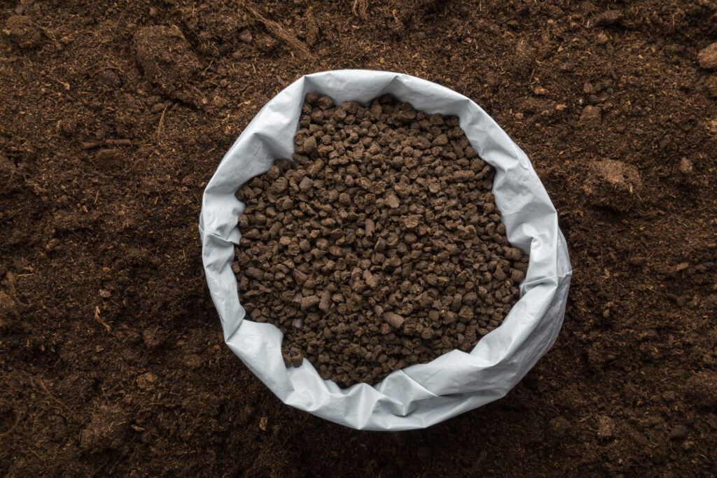 Opened plastic bag with black granules of chicken manure on dark soil background. Closeup. Product for root feeding of vegetables, flowers and plants. Top down view.