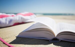 Beach scene with slight breeze and waves breaking in distance. Straw beach mat with red and white beach towel and opened book. Sun is shining on the sand and water with blue skies.