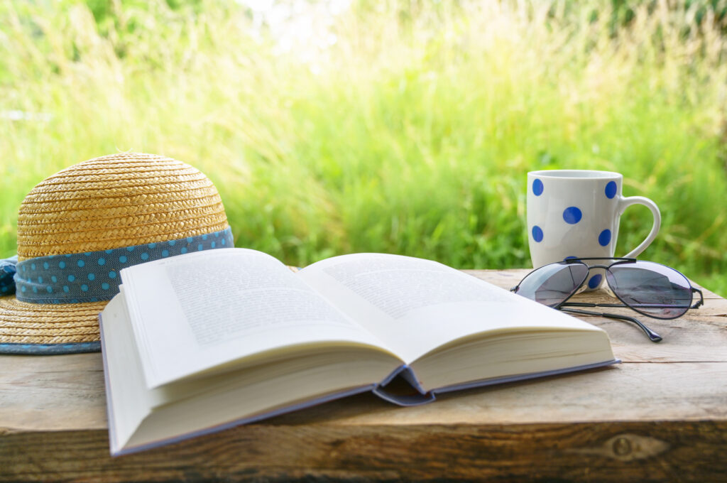 at home with book, sun glasses, coffee mug and straw hat on a rustic wooden table in the garden